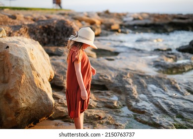child in a dress and a hat walks barefoot along the seashore, a girl walks along the water's edge, rest and tranquility on the sea coast, rocks and red stones