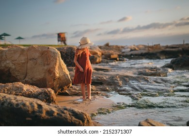 child in a dress and a hat walks barefoot along the seashore, a girl walks along the water's edge, rest and tranquility on the sea coast, rocks and red stones