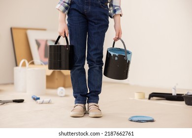 The child dressed in jeans and checked shirt holds cans of paint in hands. The girl's face is not visible, only new ones. In background are paintings, paint rollers, paint and other things for repair.