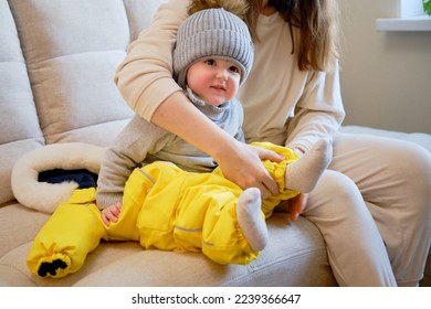 Child is dressed in warm winter clothes for a walk outside. Woman mother puts clothes on toddler baby boy for cold weather. Kid age one year five months in the home living room