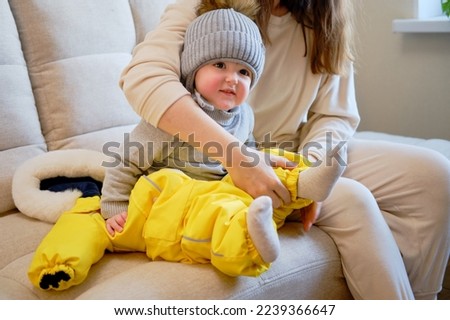 Child is dressed in warm winter clothes for a walk outside. Woman mother puts clothes on toddler baby boy for cold weather. Kid age one year five months in the home living room