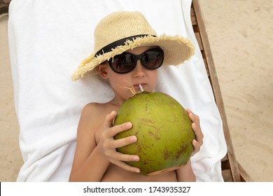 The Child Drinks Coconut Juice From A Whole Coconut On The Beach.The Cute Boy In Sunglasses And Straw Hat Rests  On A Sunbed.Image With Selective Focus.