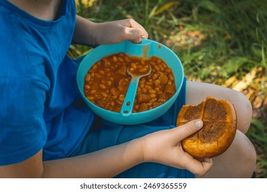 child eating a meal, baked beans, beans in tomato sauce with sausage and roll on a picnic by the lake in nature outdoors - Powered by Shutterstock