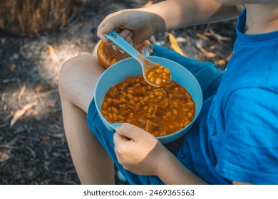 child eating a meal, baked beans, beans in tomato sauce with sausage and roll on a picnic by the lake in nature outdoors - Powered by Shutterstock