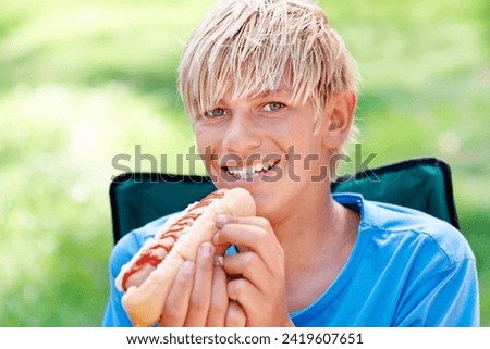 Child, eating and portrait with hotdog outdoor in camping chair and relax at barbecue with lunch. Happy, kid and hungry for food from bbq in park, woods or forest on holiday or vacation in summer