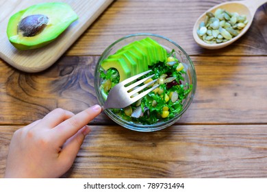 Child Is Eating A Salad. Child Is Holding A Fork In His Hand. Salad With Fresh Avocado, Lettuce Leaves, Canned Corn And Pumpkin Seeds In A Glass Bowl And On A Wooden Table. Healthy Food For Kids