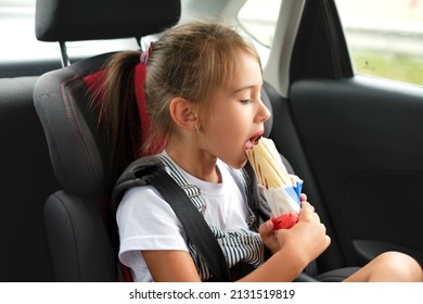 A child eats a hot dog in a car. The girl in the back seat of the automobile is fastened with seat belts during the trip. Takeaway food