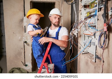 Child electrician standing in ladder and holding pliers tool while installing switchboard with father at home. Man and kid mounting electrical wiring in apartment under renovation. - Powered by Shutterstock