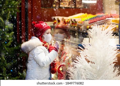 Child In Face Mask At Store Window On Traditional Christmas Market In Coronavirus Outbreak. Covid-19 Pandemic. Safe Xmas Shopping, Gifts And Presents With Kids On Snowy Winter Day. 