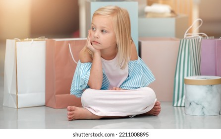 Child, Fashion And Shopping Bags While Looking Bored And Waiting In A Kids Clothing Shop, Store Or Boutique With Cute Girl Sitting On Floor. Little Kid Shopper Or Customer With Sale Purchase Packages