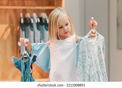 Child, fashion and shopping while choosing kids clothing in a shop, store or boutique with a cute girl holding dresses thinking or picking color choice. Little kid shopper or customer buying clothes
