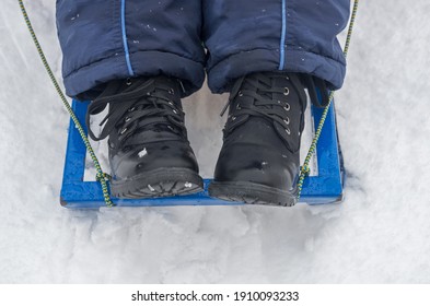 Child Feet In Blue Winter Pants And Black Lace-up Shoes, Dusted With Snowflakes, Stand On Edge Of Sled With Blue Metal Frame And Runners With Tight Rope Rein Attached. Sleigh Is On Fluffy Snowdrift.
