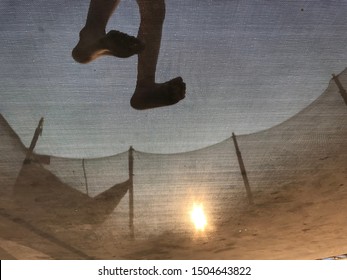 Child Feet Seen From Below A Trampoline Against The Setting Sun.