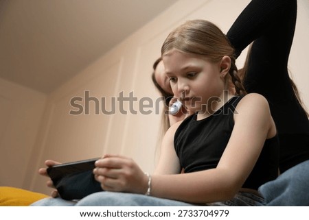 Child focused on smartphone while mother watches nearby, illustrating family screen time, modern parenting and digital lifestyle at home with bonding and casual interaction