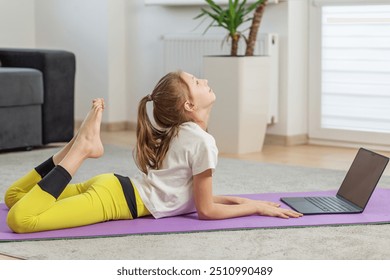 Child following an online yoga class while performing backbend on yoga mat at home, focusing on flexibility and posture during practice. - Powered by Shutterstock