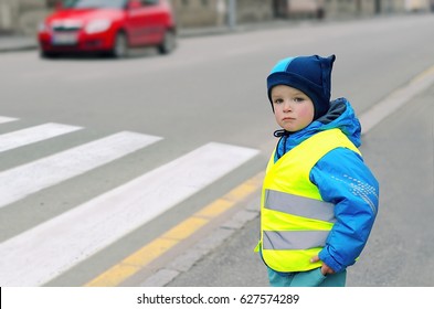 Child In Front Of Pedestrian Crossing. Little Boy Finds Out If He Can Cross The Zebra Crossing. He Wears Reflective Vest Because Of Safety. Car In The Background. Child Concept. Traffic Concept.