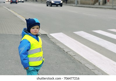 Child In Front Of Pedestrian Crossing. Little Boy Finds Out If He Can Cross The Crossswalk. He Wears Reflective Vest Because Of Safety. Car In The Background. Child Concept. Traffic Concept.