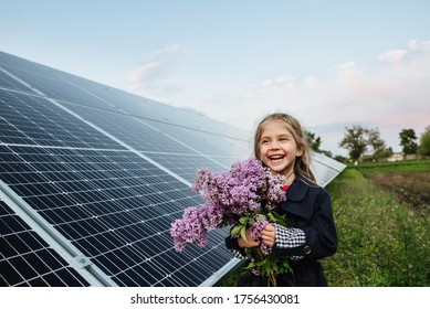 A Child With A Future Of Alternative Energy And Sustainable Energy. The Child Holds Flowers On A Background Of Solar Panels, Photovoltaic. Environmental Friendliness And Clean Energy Concept.