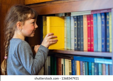 The Child Gets A Textbook With Wooden Shelf Of The Old Library. A Little Girl Is Taking A Yellow Book From A Bookshelf. Children's Educational Concept.