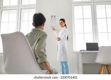 Child Getting Eye Checkup At Doctor's Office. Friendly Ophthalmologist Standing In Modern Medical Office, Doing Eye Exam, Testing Vision Of Little School Boy And Pointing At Eye Chart On White Wall