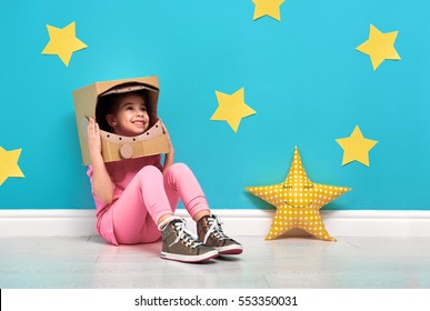 Child Girl In An Astronaut Costume Is Playing And Dreaming Of Becoming A Spacemen. Portrait Of Funny Kid On A Background Of Bright Blue Wall With Yellow Stars.