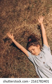 Child Girl In A Denim Dress Lies On A Haystack Stretching Her Arms Up. Holidays At Grandma's In The Village