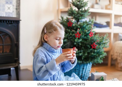 Child Girl Is Drinking Hot Beverages By Christmas Tree At Home. Kid Holds Mug With Steamy Tea Or Cocoa Near Wood Burning Stove In Cozy Warm Living Room. Concept Of Winter Mood, Cold, Chilly, New Year