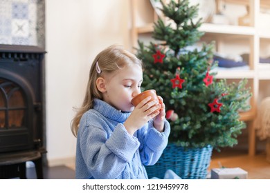 Child Girl Is Drinking Hot Beverages By Christmas Tree At Home. Kid Holds Mug With Tea Or Cocoa Near Wood Burning Stove In Cozy Warm Living Room. Concept Of Winter Mood, Cold And Chilly, New Year