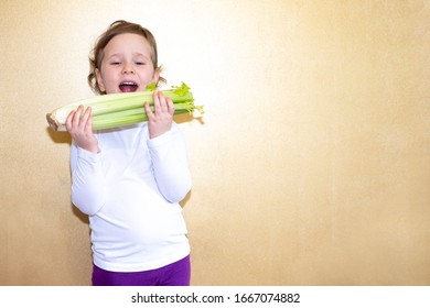 A Child, A Girl, A European Holds Celery In His Hands. Girl In A White Jumper On A Yellow Background With Vegetables In Her Hands.