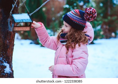 Child Girl Feeding Birds In Winter. Bird Feeder In Snowy Garden, Helping Birds During Cold Season, Teaching Kids To Love And Protect Nature