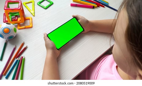 Child Girl Holding Smartphone With Green Screen Horizontal Position On White Wooden Table Background. Online Education Concept. Mobile Phone With Chroma Key. Close Up Hands.