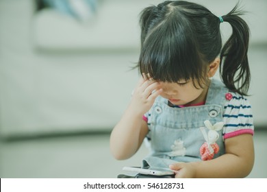 Child Girl Look Smart Phone And Holding Her Head With Hands Trying To Remember Something Or Having Headache.Child Development Concept.Little Asian Girl Look Smart Phone In Living Room And Headache.