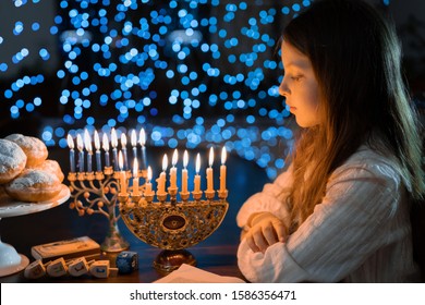 Child Girl Looking At Menorah Candles On Wooden Table And Sufganiyot On Background Light Glitter Bokeh Overlay. Hanukkah Jewish Holiday Israel Hebrew Traditional Family Celebration Invitation Design