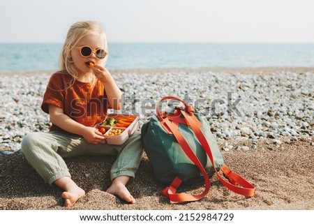 Child girl with lunchbox eating vegetables outdoor travel vacation healthy lifestyle vegan food picnic on beach hungry kid with lunch box snacks and backpack