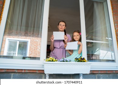 A child girl with mother woman from the window shows a white blank sheet for a message with an copy space during the home isolation from the coronavirus pandemic - Shutterstock ID 1707440911