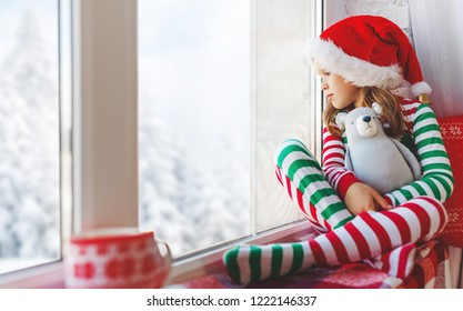 Child  Girl In Pajamas Is Sad On Christmas Morning By The Window
