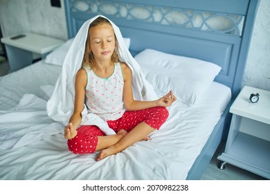 Child girl in pajamas sitting on a bed in lotus pose, Little girl wakes up from sleep on a big and cozy bed white linen in the at home, family lifestyle, comfortable bed, Good morning