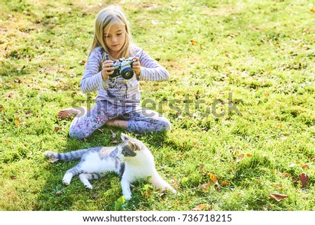 Child girl photographing a cat lying on the grass outside in the garden. Focused to cat