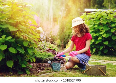 Child Girl Playing Little Gardener And Helping In Summer Garden, Wearing Hat And Gloves, Working With Tools