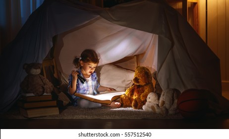 child girl reading  with  book and flashlight and teddy bear in tent. before going to bed