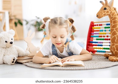 a child girl reads a book lying on the floor among toys in a bright bright nursery or kindergarten, the concept of children's learning centers or kindergartens and learning. - Powered by Shutterstock