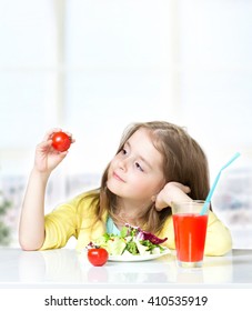 Child Girl Sitting At Table Eating Fresh Salad Drinking Juice. Female Kid Healthy Nutrition Concept Background.Toddler Having Lunch Holding Tomatoes In Hands.