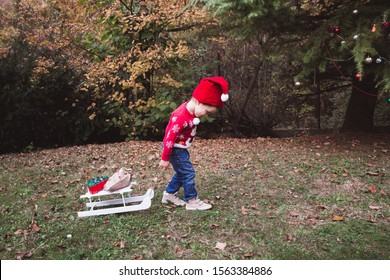 Child Girl Is Smiling Under Christmas Tree With Red Ornaments Outside. Little Kid In Santa Hat Bring Presents On Sled