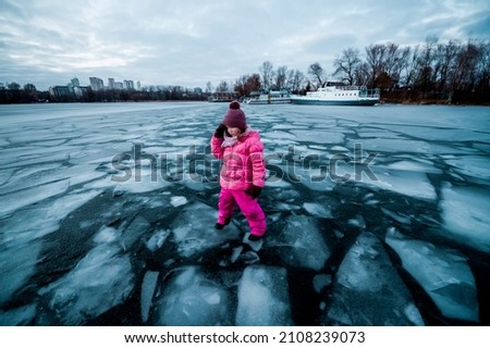 Child girl is standing on cracked ice. skating on frozen canal with snow. Little girl with skates on natural ice on cold winter days. Kid playing on ice with ship on background. landscape.