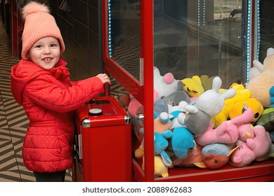 A Child Girl Stands Near A Machine With Soft Toys And Plays, Wants To Catch And Pull Out A Toy Prize