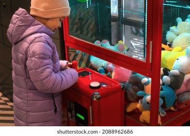 A Child Girl Stands Near A Machine With Soft Toys And Plays, Wants To Catch And Pull Out A Toy Prize