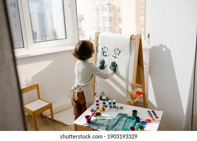 Child Girl Wearing Apron Painting Making Fingerprints On Paper On Easel In Children Room. Finger Painting Or Art Therapy For Children. Fun Activities For Toddlers.