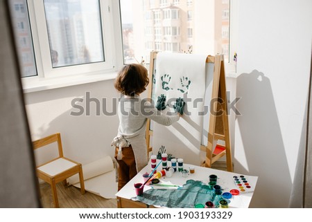 Child girl wearing apron painting making fingerprints on paper on easel in children room. Finger painting or art therapy for children. Fun activities for toddlers.
