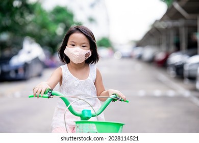 Child Girl Wearing Cloth Face Mask Driving Green Bicycle At Outdoors. Preventing Spread Coronavirus (covid-19) Outbreak Infections. Happy Kid Aged 4-5 Years Old.