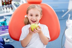 Child Girl In White Clothes At Reception At Pediatric Dentist Smiles And Laughs. Little Girl Is Holding A Big Green Apple In Her Hands. Concept Is Children's Health.